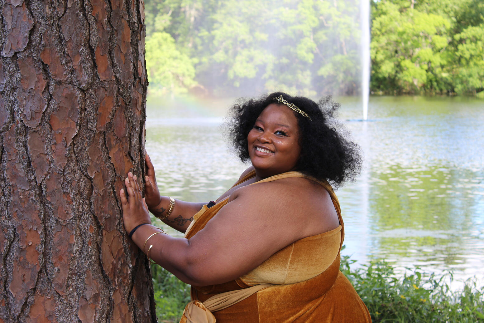 Golden goddess posing on a tree in front of a lake surrounded by green trees.