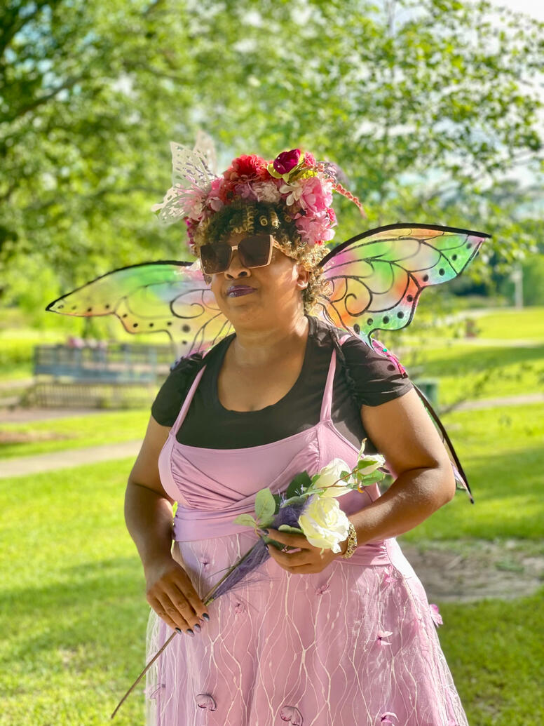 fae with authority looking forward with large shades over eyes and flower wand in hand.