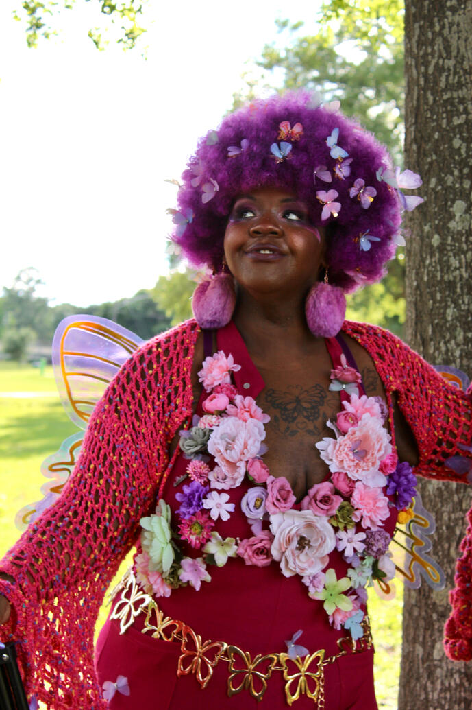 black fae mobile host looking towards the sky and facing the camera with purple afro and adorned all over with flowers and butterflies.