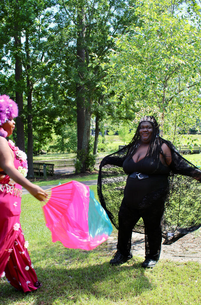 Magenta fae fanning dark fae who poses regally with her lace cape spread amongst the trees.